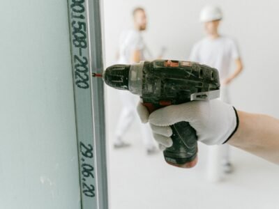 Mesin Bor Impact Cordless - A construction worker uses a drill on drywall with blurred workers in the background.
