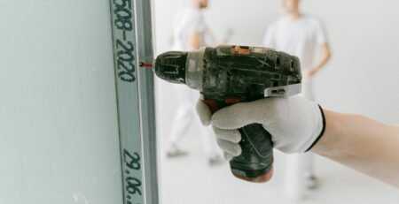Mesin Bor Impact Cordless - A construction worker uses a drill on drywall with blurred workers in the background.