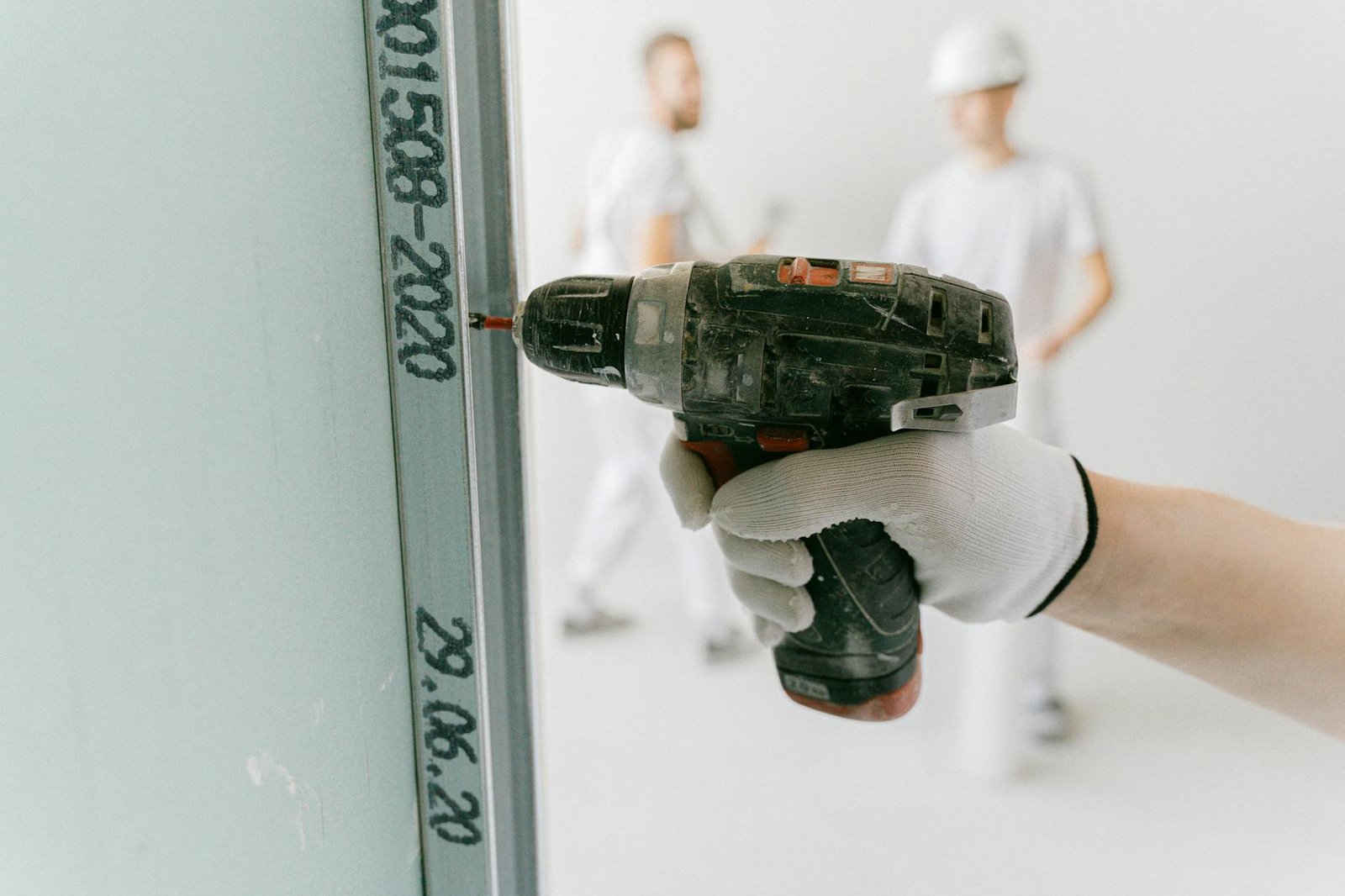 Mesin Bor Impact Cordless - A construction worker uses a drill on drywall with blurred workers in the background.