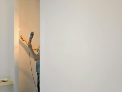 Mesin Bor Impact Cordless - Unrecognizable person using a drill on a minimalist white wall in a home interior.
