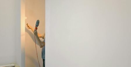 Mesin Bor Impact Cordless - Unrecognizable person using a drill on a minimalist white wall in a home interior.