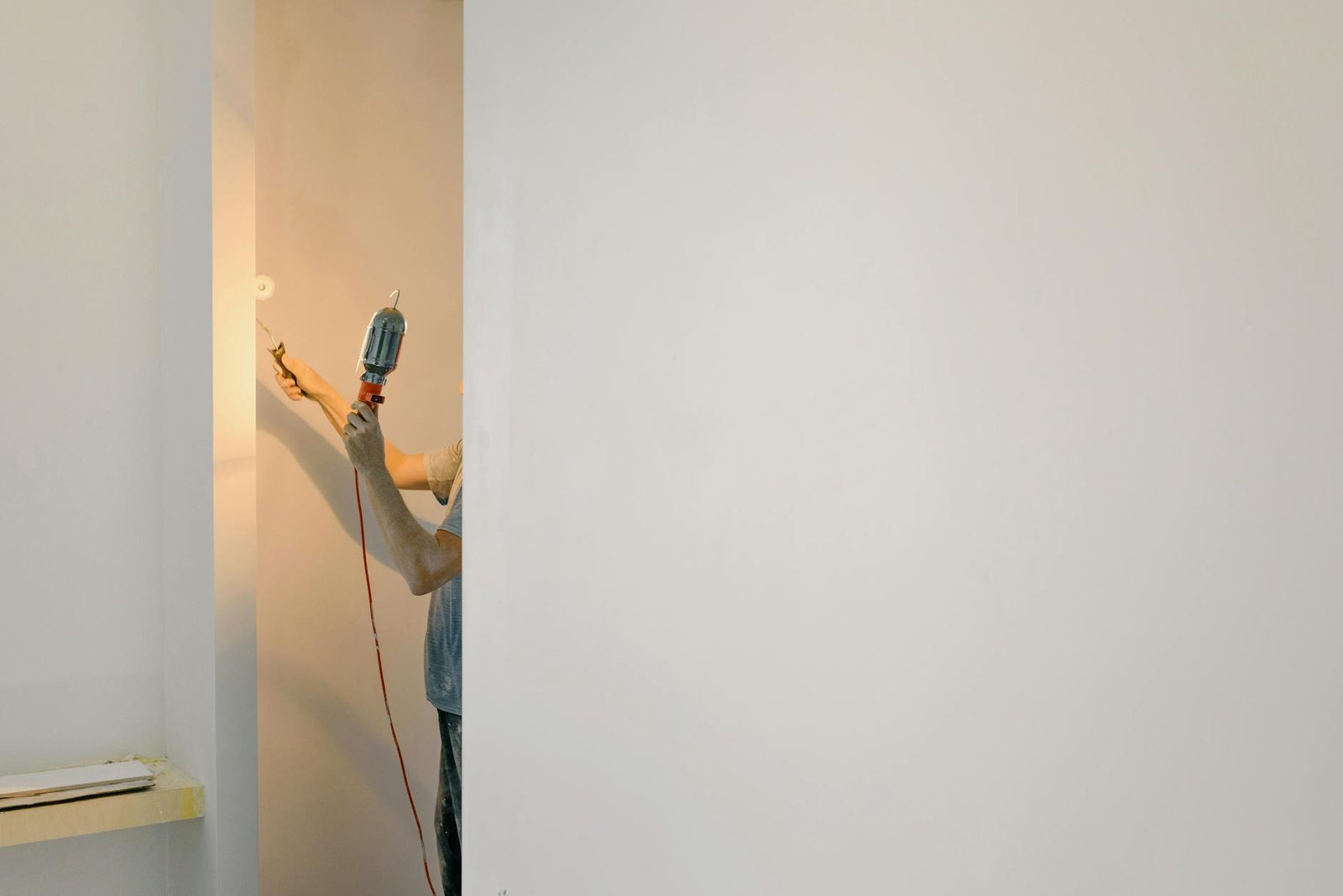 Mesin Bor Impact Cordless - Unrecognizable person using a drill on a minimalist white wall in a home interior.
